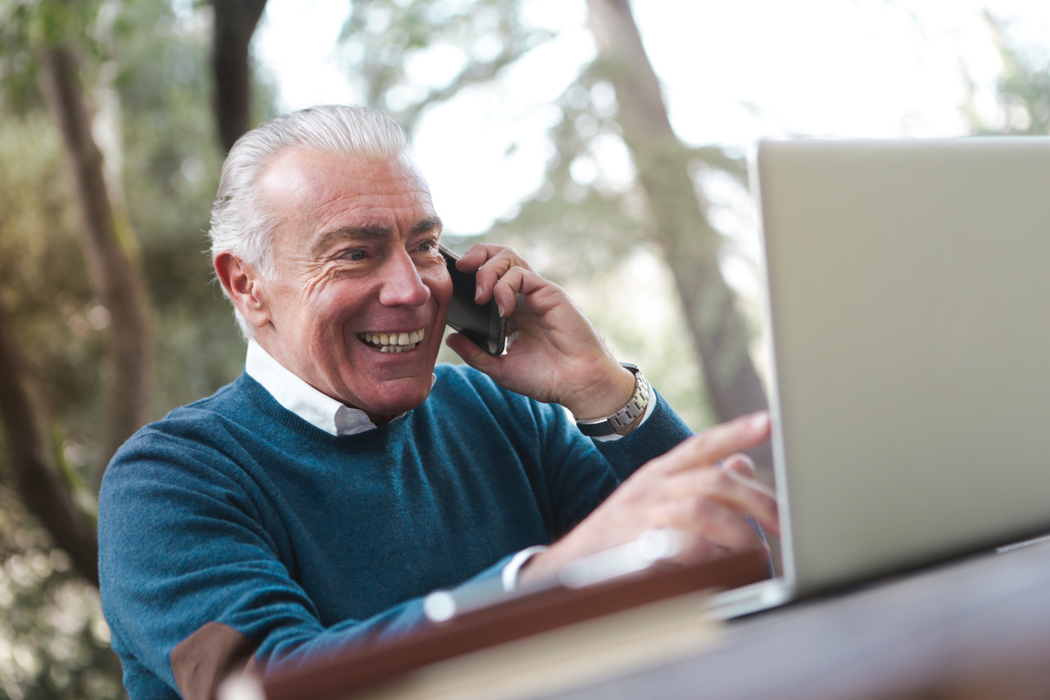 Selective Focus Photo of Smiling Elderly Man in Blue Sweater Sitting by the Table Talking on the Phone While Using a Laptop